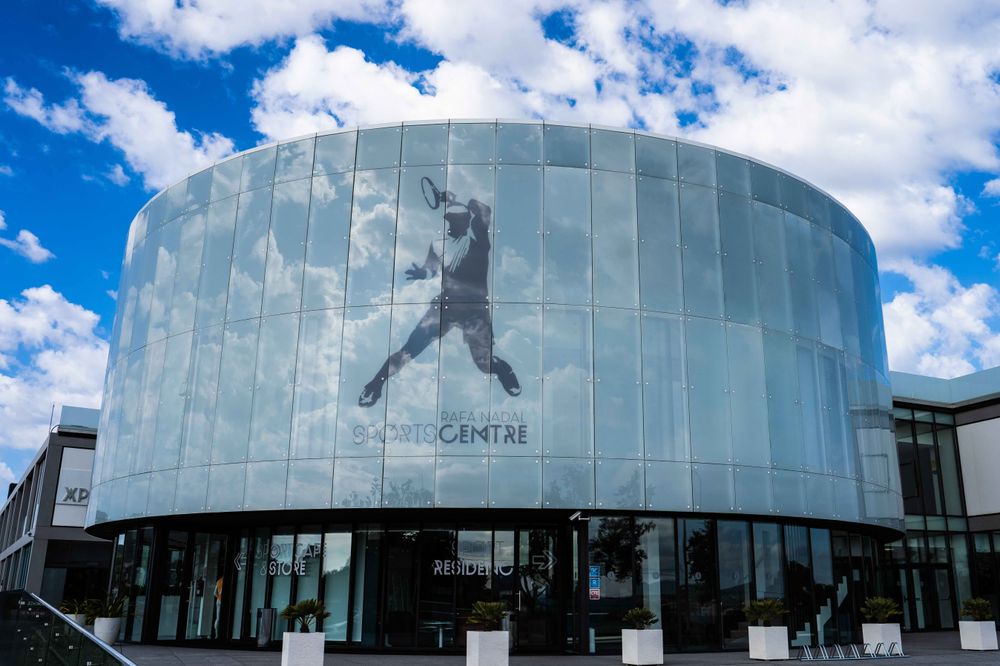 A modern building with a cylindrical glass facade featuring a silhouette of a tennis player in action, likely Rafael Nadal, against a partly cloudy sky.