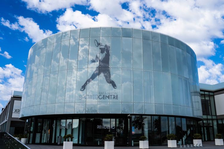 A modern building with a cylindrical glass facade featuring a silhouette of a tennis player in action, likely Rafael Nadal, against a partly cloudy sky.